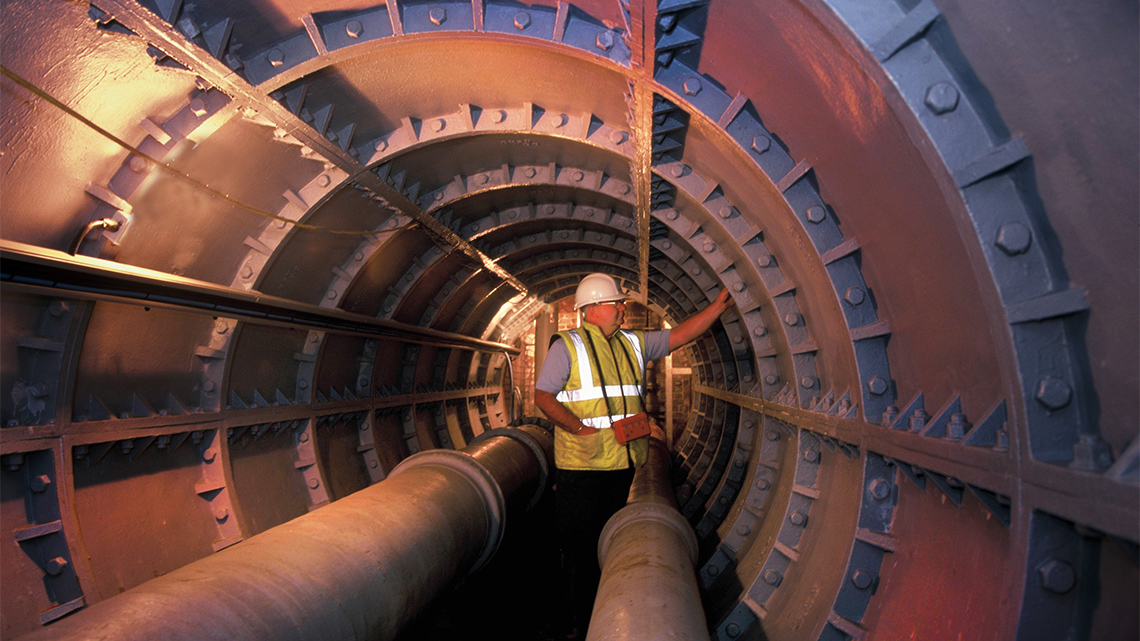 A worker inspecting a metal tunnel with two large water pipes running through it.