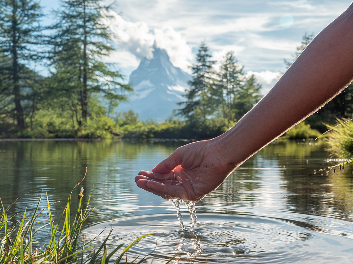 A hand scooping clean water our of a lake surrounded by trees with mountains in the background.