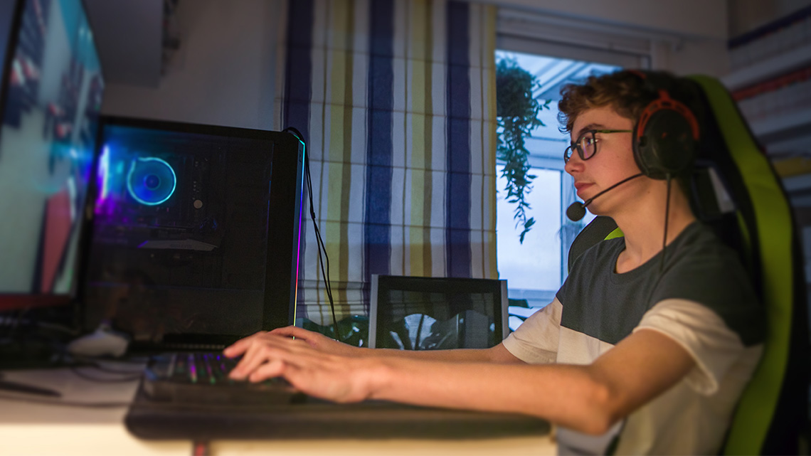 A teen is sitting in their comfortable desk chair, wearing headphones and typing on a keyboard.