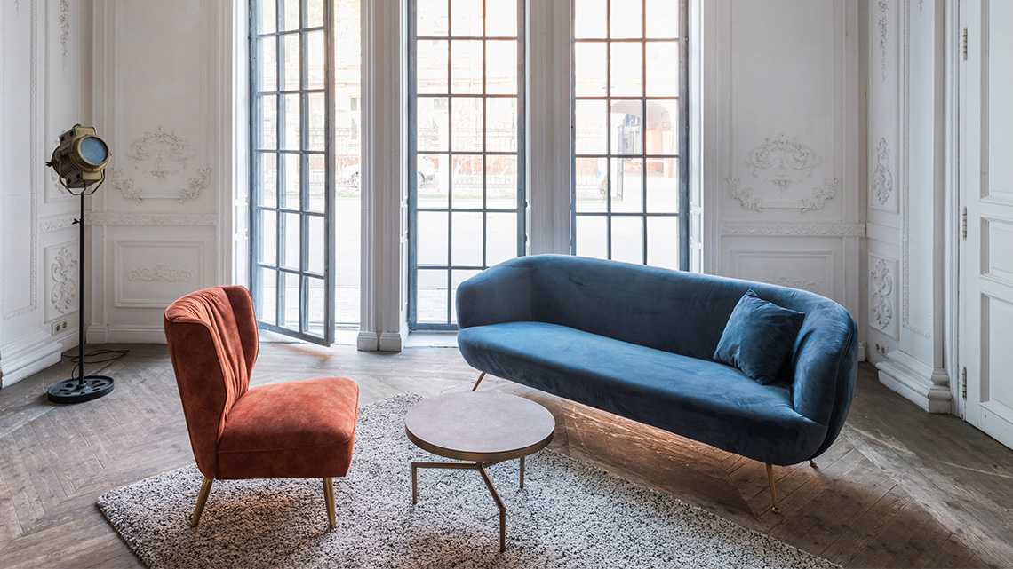 A chair and a sofa with a small round table between them. Behind the sofa is a set of windows and a tall lamp.