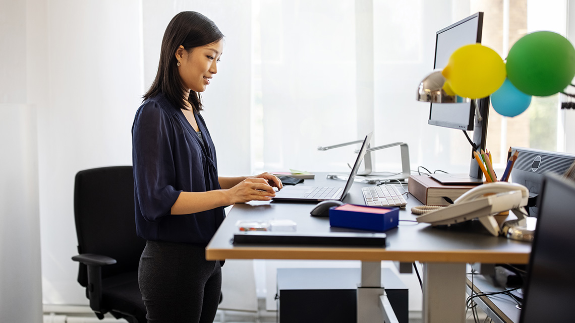 A person standing and working at a raised desk. She works on a laptop with an additional monitor. On her desk is a mouse, keyboard, telephone, and papers.