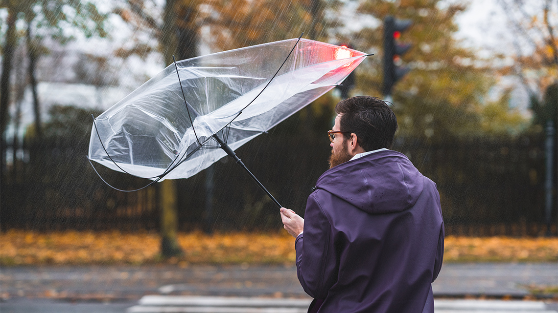 A person standing in the rain holding an umbrella that is being blown inside-out by strong winds.