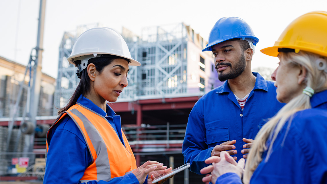 A group of civil engineers, wearing hard hats and construction jumpsuits, discuss plans outside a building under construction.