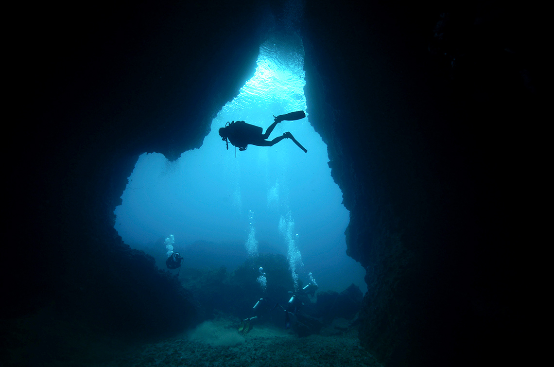 Silhouettes of divers in an underwater cave with light shining down from the surface.