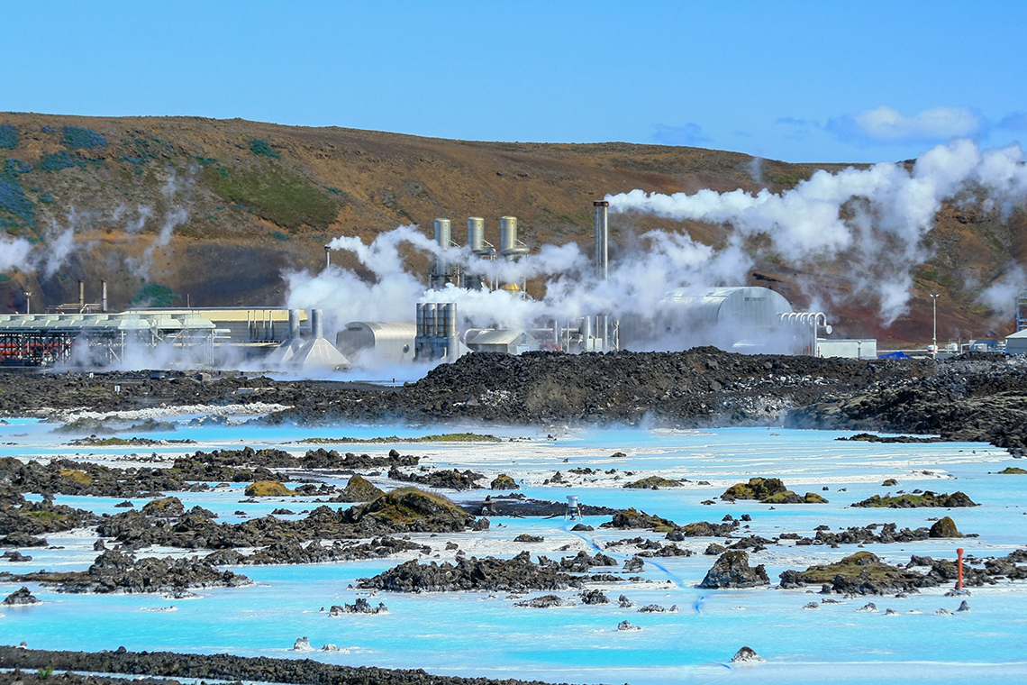 A geothermal power plant with steam coming out of several chimneys next to a steaming geyser.