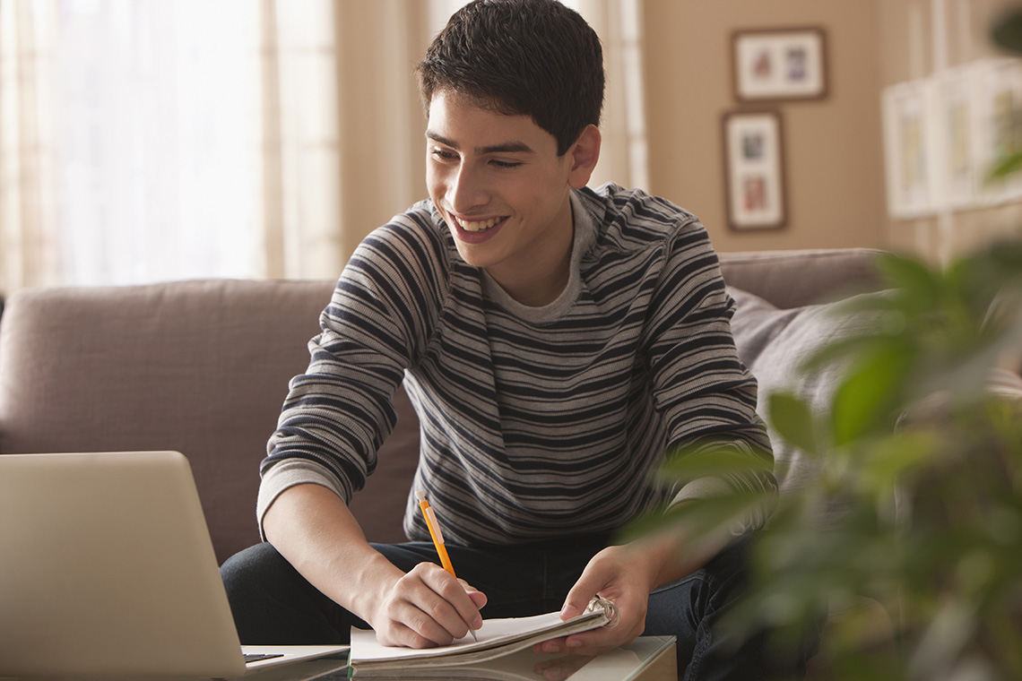 A person smiling at the computer while writing in their notebook.