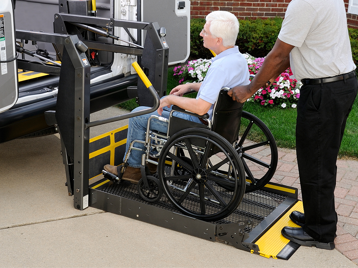 A person assists a person in a wheelchair to use an elevator ramp to enter a bus.