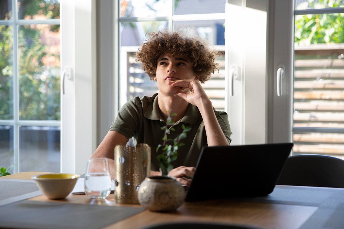 A person sitting at a table with their laptop open and thinking.