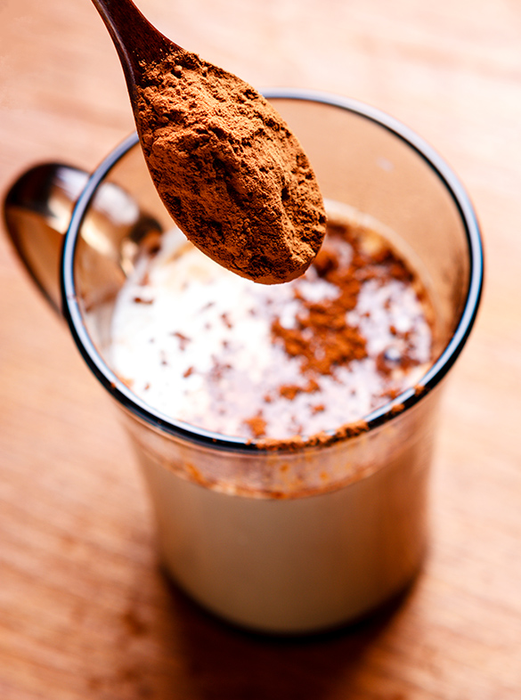 A spoon of chocolate milk powder is being added to a glass of milk.