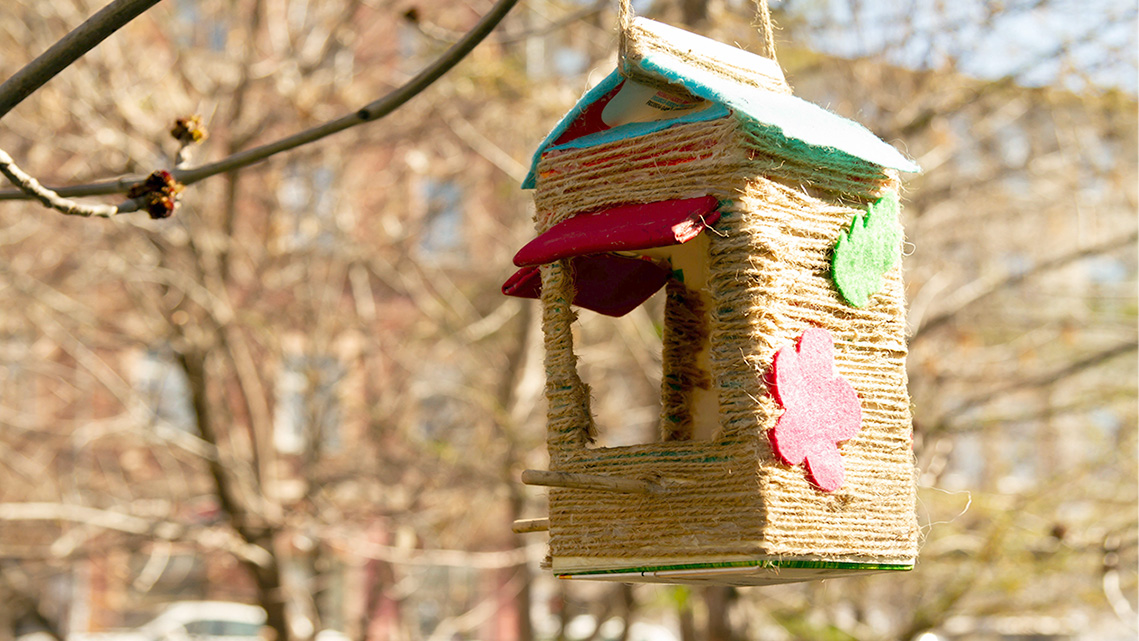 A milk carton covered in twine hangs from a tree.  There are two perches for birds to land and two holes in the carton where bird feed is piled.