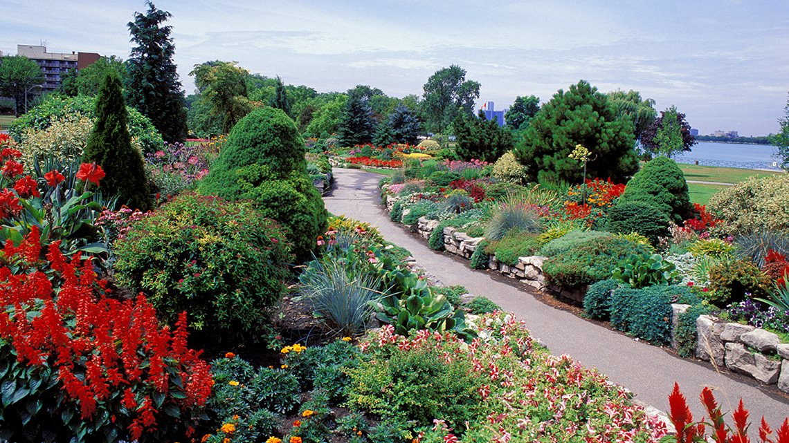 A variety of flowers, bushes, and trees line a park path near a river.