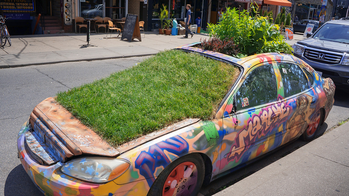 A car with plants growing out of its hood, windshield, sunroof, and trunk next to a sidewalk.