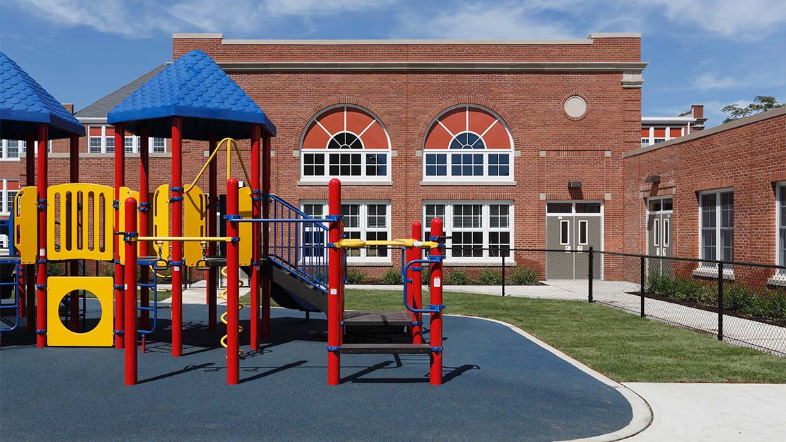 A playground with stairs, monkey bars, a tunnel, and a bridge next to a school.