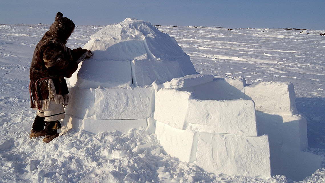 An Inuit person building a small dome-shaped structure made of blocks of frozen snow.