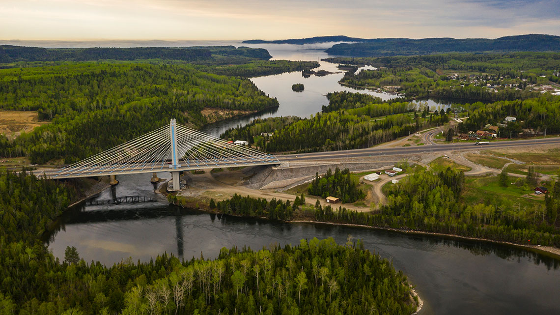 There is a large suspension bridge over a narrow, winding river surrounded by trees.