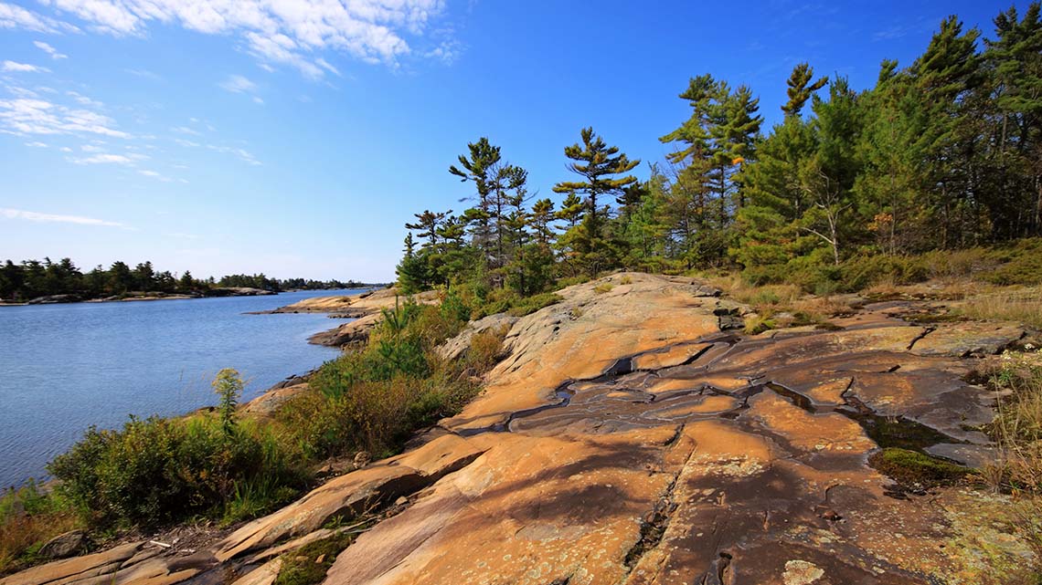 A Canadian Shield rock face shown along the shore of a narrow lake on a sunny day. An island with trees is on the far side of the lake.