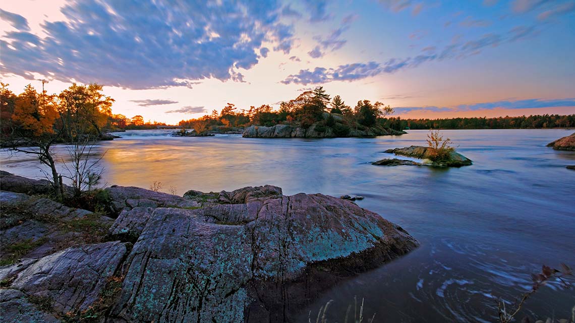 A lake with a rocky Canadian Shield shoreline has several small islands made of rock, with plants and trees growing on the rocky islands.