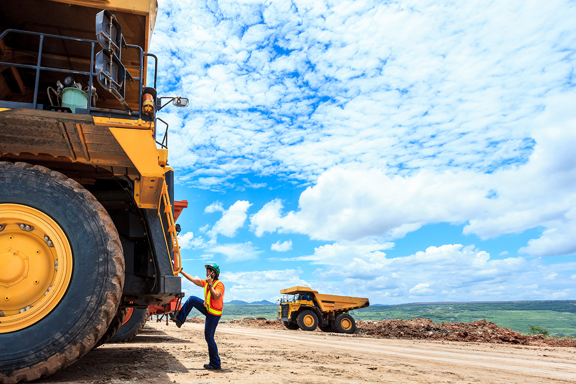 A person wearing a hard hat and construction vest, and talking into a walkie talkie radio, climbs up onto the back of a huge mining machine.