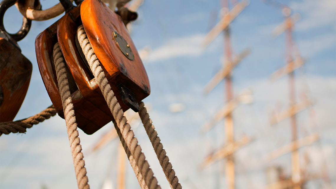 A close-up view of a wooden block and tackle pulley system with a rope through it, in front of a blurred background of tall ship masts.