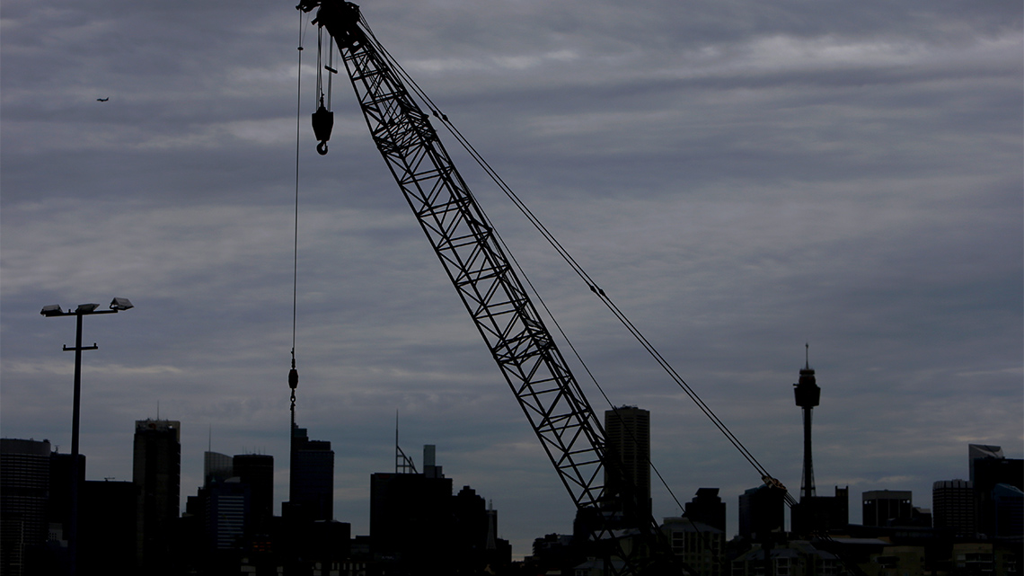 A city skyline shown at twilight as the sky is darkening with a silhouette of a large construction crane in the foreground.