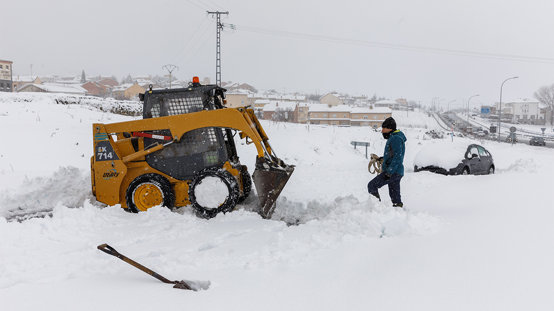 A man dressed in winter clothing stands in the snow outdoors in front of a skid steer that is stopped after plowing a portion of a sidewalk.