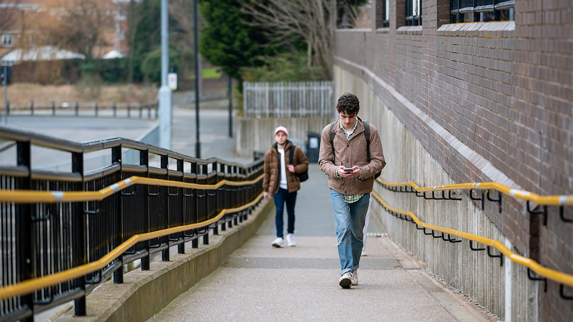 Two young people walk towards you up a concrete ramp alongside a building outdoors. The ramp has strong railings on either side.