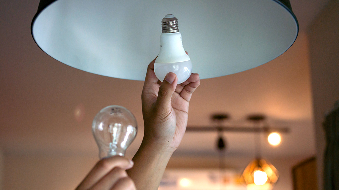 A person reaches up to insert an LED light bulb into an indoor ceiling light fixture while holding a regular light bulb in their other hand.