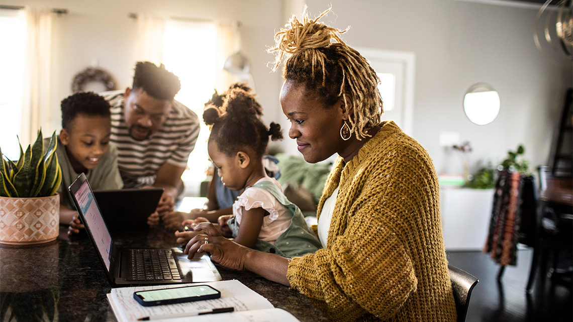 Four people sit at a table in a dining room indoors. Two people are using a tablet. One person is using a phone. One person uses a laptop.