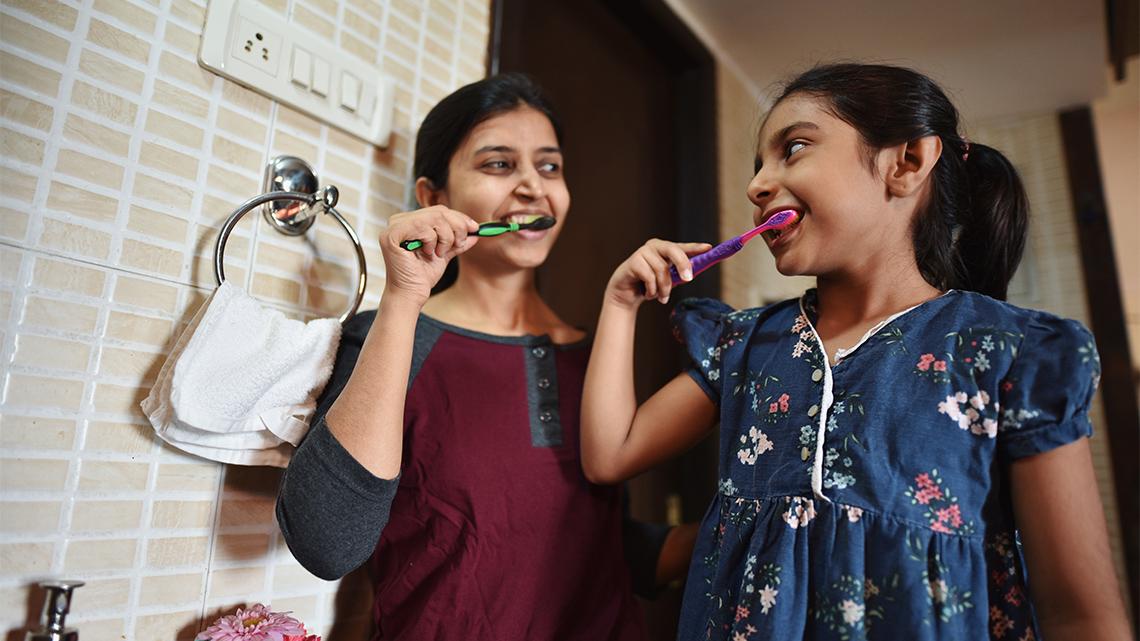 Two young people stand in a bathroom indoors, looking at each other as they brush their teeth using toothbrushes with toothpaste.