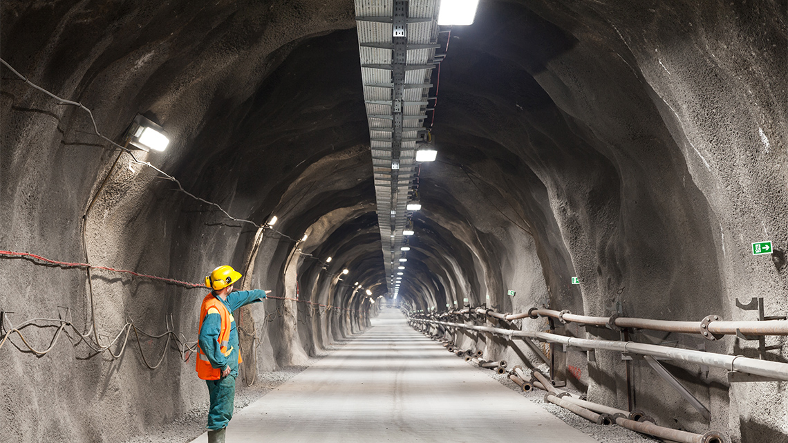 An underground mining tunnel has lights at the top and sides, with pipes on one side. A mine worker points down the tunnel away from you.