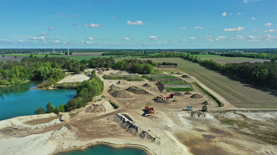A large area of land and water that is being cleared and dug for mining by large vehicles. Three wind turbines are seen off in the distance.