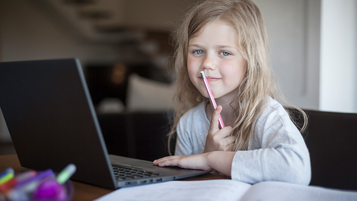 A young person sits at a desk inside in a well-lit room in front of a laptop holding an eraser-topped pencil in front of their closed mouth.