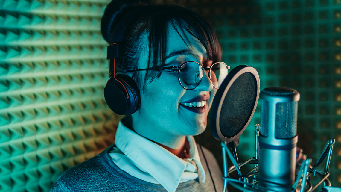 A woman wearing headphones sings into a microphone in a soundproof room. The walls of the room are covered in sound-absorbing material.