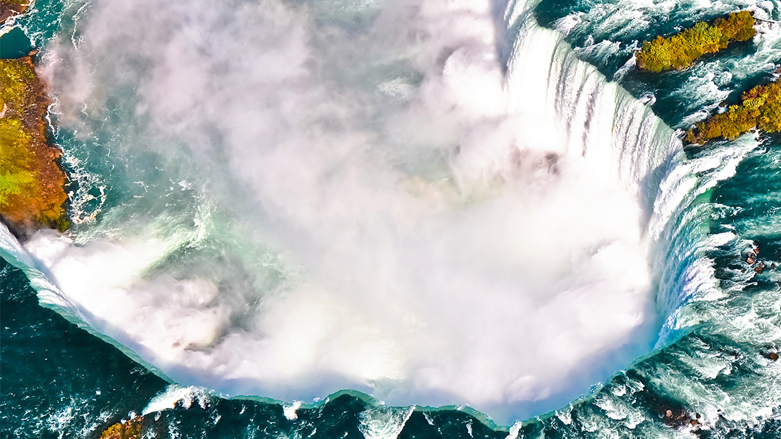 An aerial view of Niagara Falls. Water foams as it rushes past the edge and creates foam as it falls.