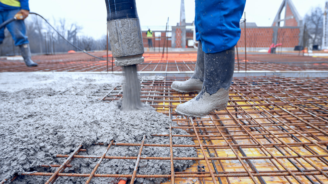 A person pouring cement through tube onto a roof. The roof has a base of wood and then a grid of metal pipes across it.