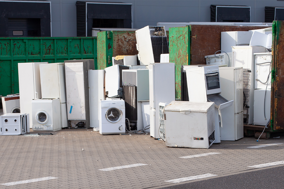 A pile of old electrical household appliances outside by a dumpster. These appliances include a freezer, microwave, fridge, and a stove top.