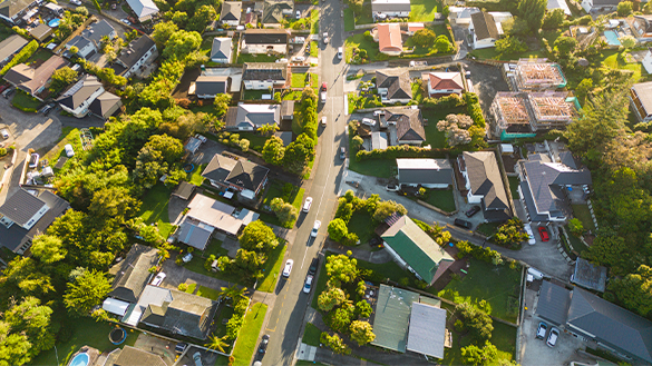 A drone view of a neighbourhood. There are houses and trees. There is also a road that has cars driving along on them.