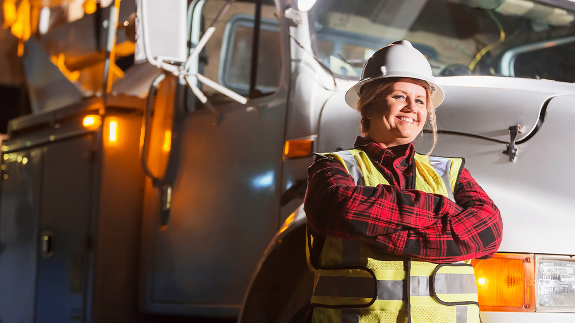 A technician is standing in front of a truck, wearing a hardhat and safety vest.