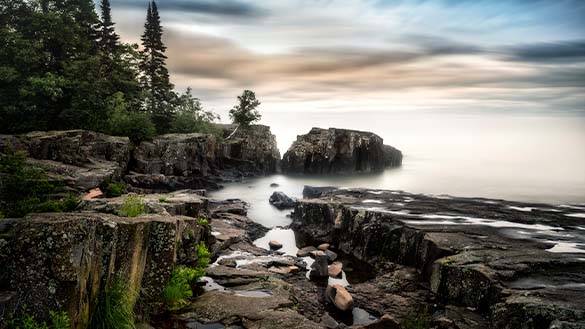 Coast of Lake Superior with large rocks and tall trees along the water.