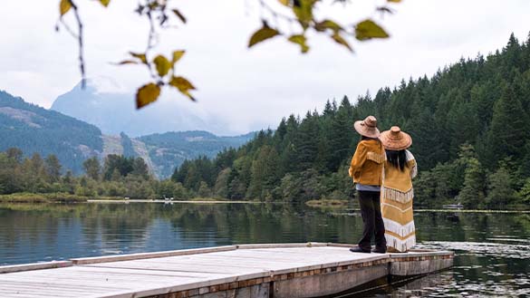 Two people standing at the edge of a dock looking out towards the water and the mountains.