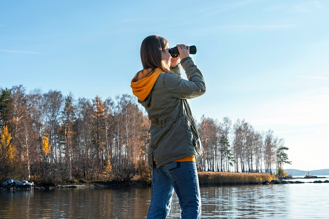 A person looking through binoculars into a lake beside a coastline of trees.