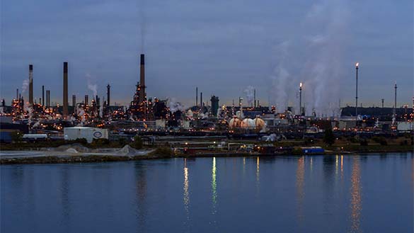 By Lake Ontario there is an oil refinery with many smokestacks with smoke rising into the air.