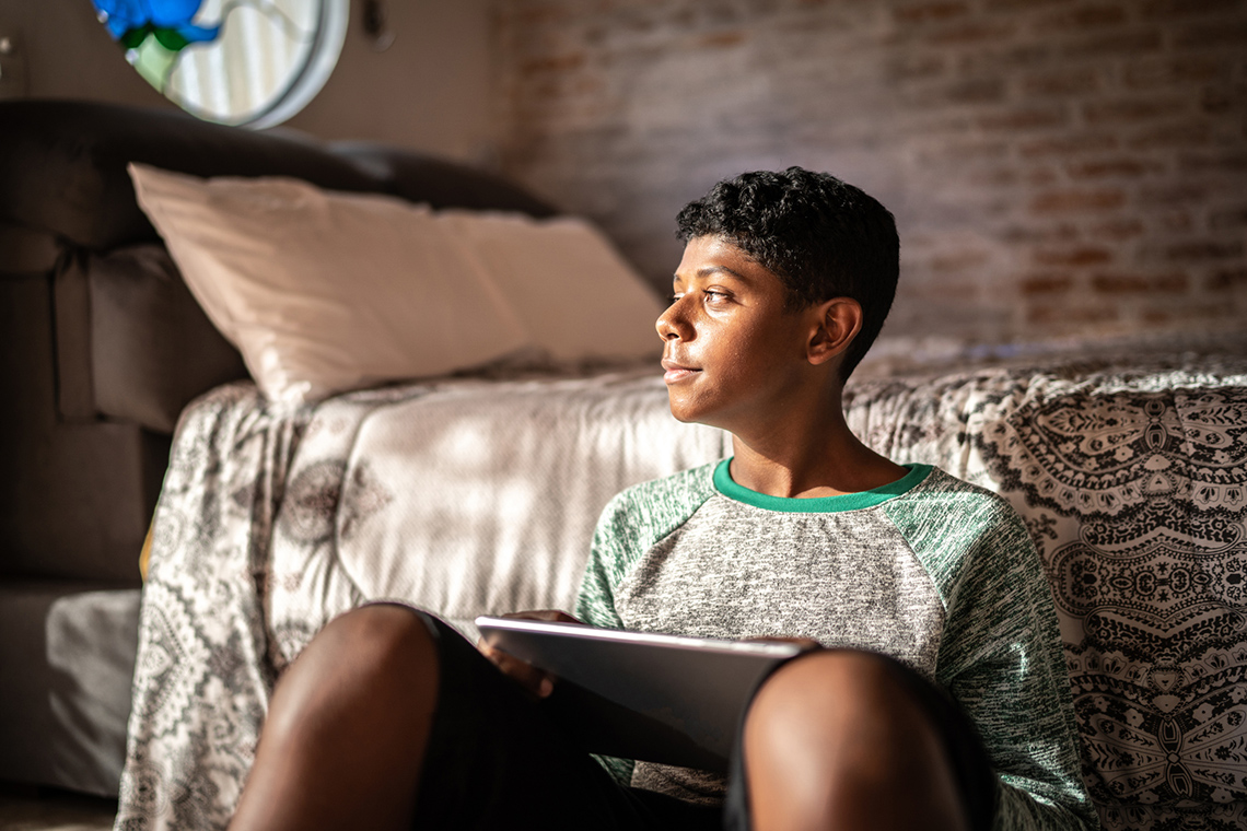 A person sitting on the floor in front of a bed holding a laptop and looking towards the window.