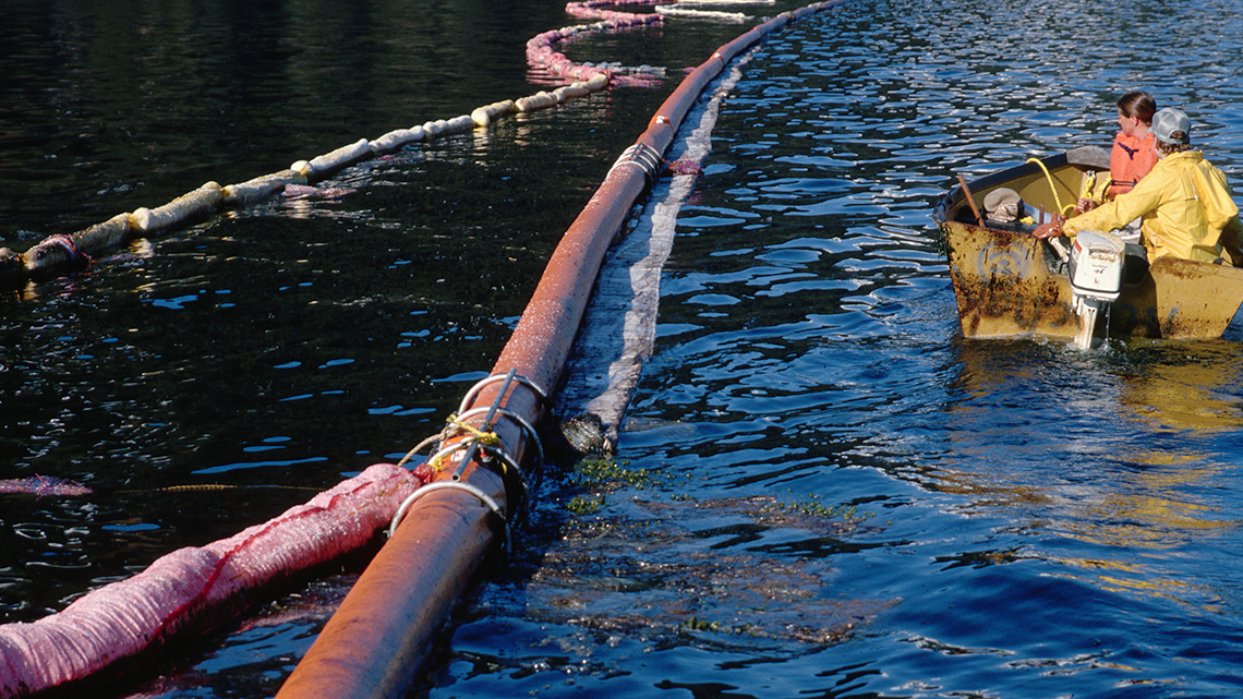 People riding in a small boat next to a pipe and sorbent materials. 