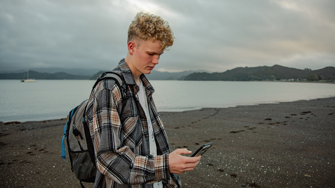A person looking at their phone while standing on the sand with a large body of water behind them.