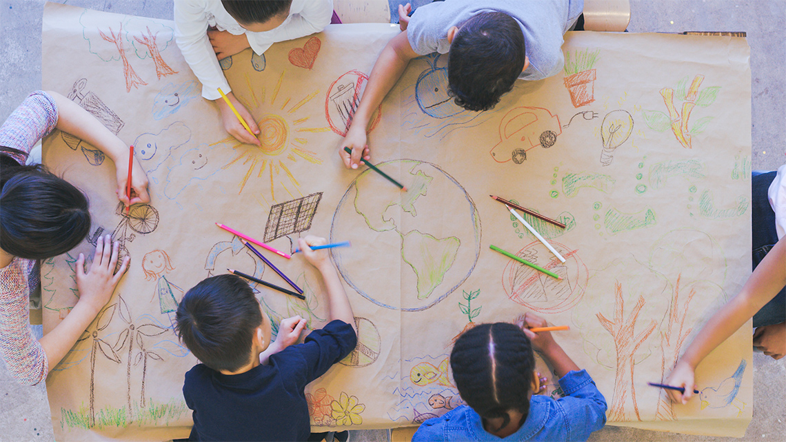  Six children are drawing a picture together on a large sheet of paper.