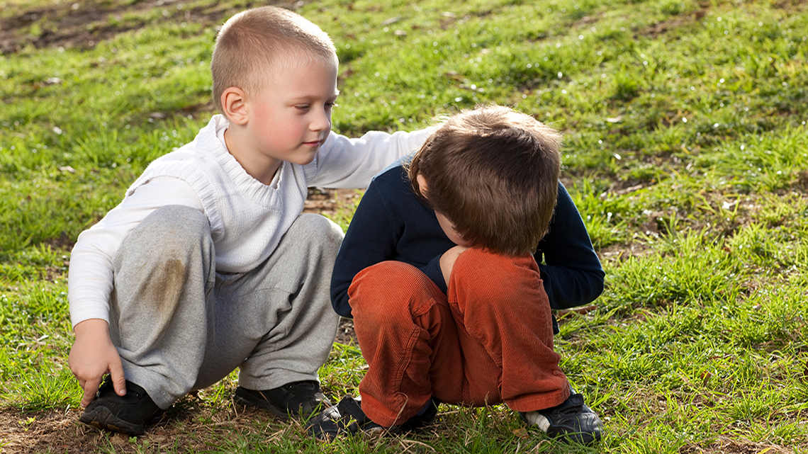 Two children sitting next to each other on the grass. One child has their hands on the other child’s back to comfort them.