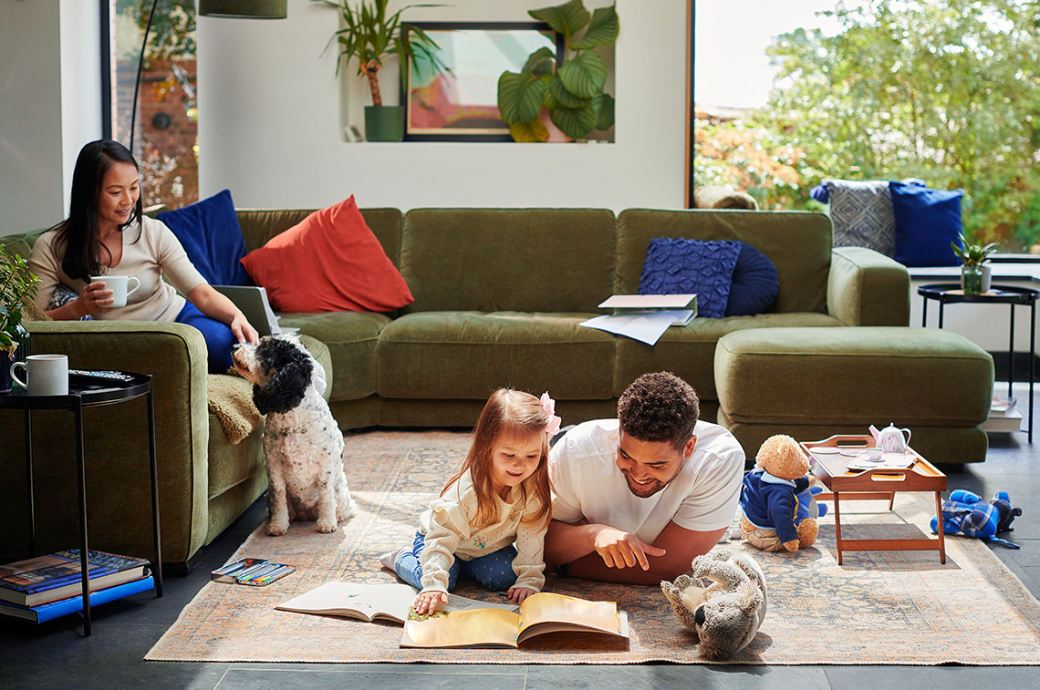 A child and an adult read a book together on a living room floor while another adult sits nearby with a dog.