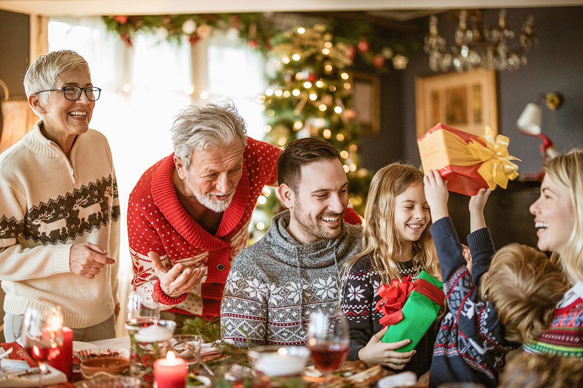 Family of 2 parents 2 grandparents & 2 kids having Christmas dinner. A Christmas tree is behind them.