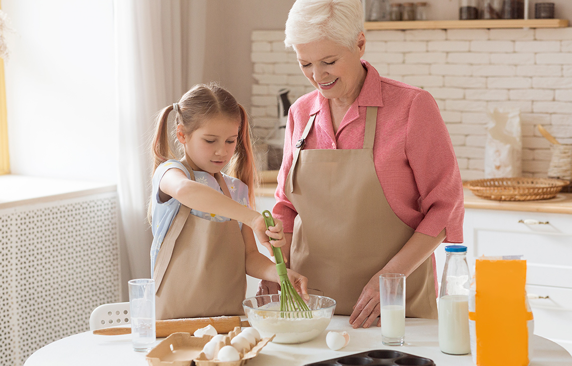 Grandma helps her granddaughter cook a special meal. Grandma watches and guides granddaughter.